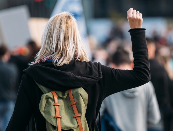 Blonde vrouw met groene rugzak houdt haar vuist omhoog op een demonstratie.